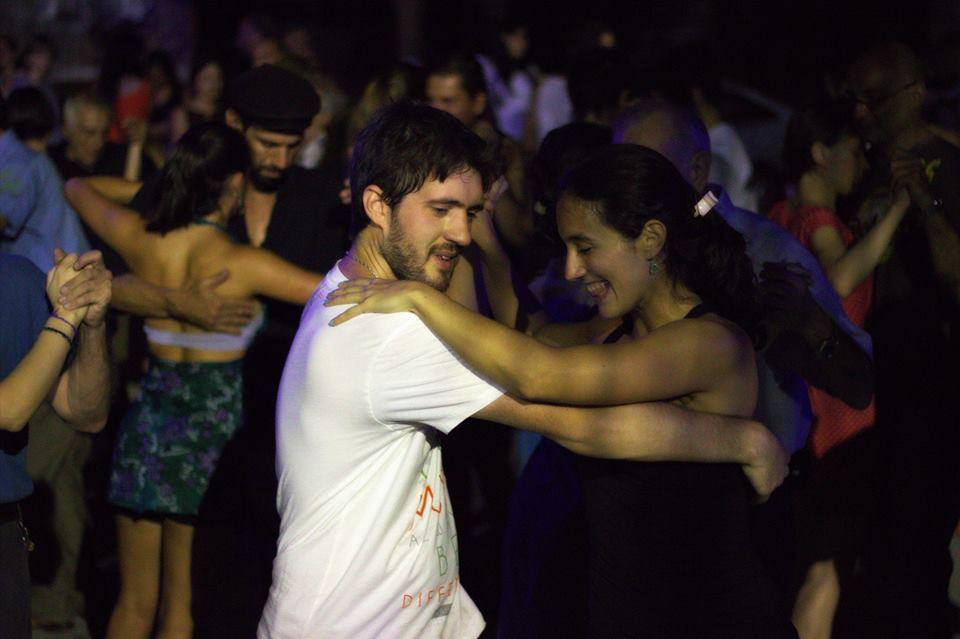 Varias parejas bailando tango en un espacio abierto. En primer plano, una pareja joven bailan mientras sonríen
