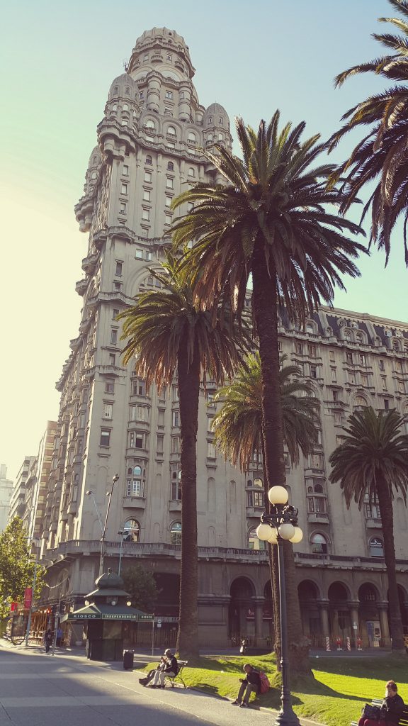 Imagen del Palacio Salvo de Montevideo visto desde la Plaza Independencia; sentados en el césped y bancos de la plaza hay varias personas.