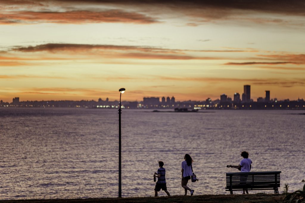 Imagen de la Rambla de Montevideo al atardecer.; tres jóvenes caminan hacia el mar: uno de ellos con un termo en la mano y otro con una guitarra.; al fondo se observa la silueta de la ciudad