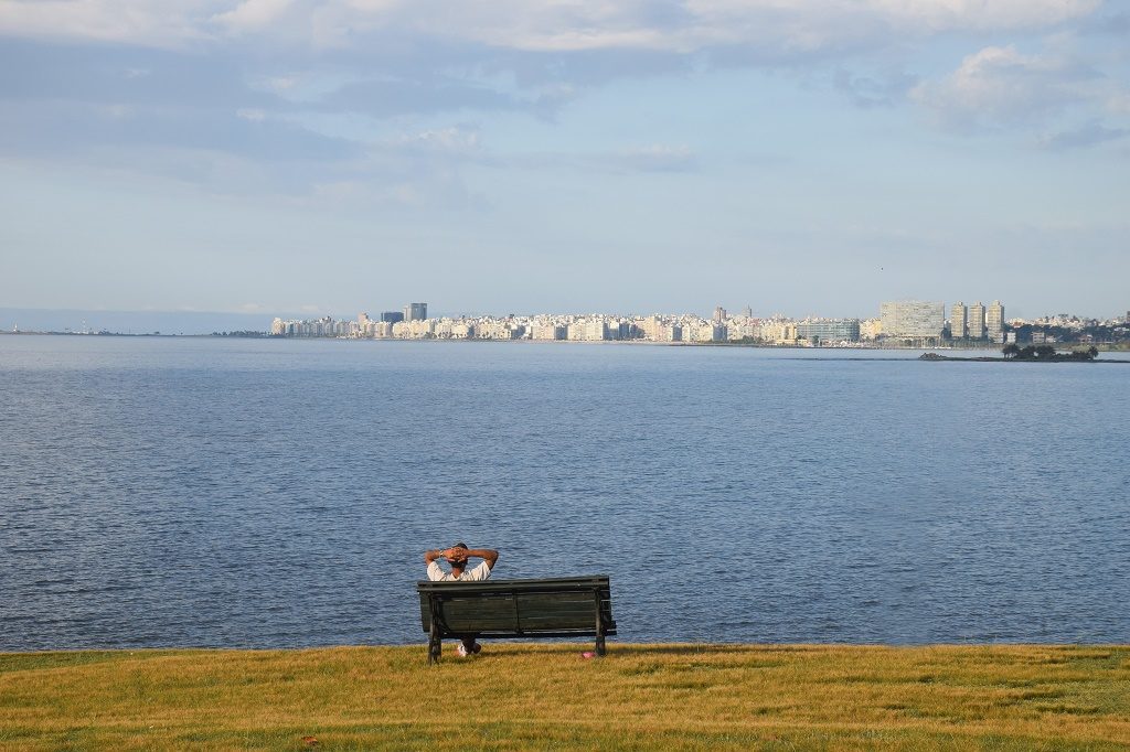 Jóven con camiseta blanca y los brazos detrás de la cabeza sentado en un banco de plaza verde sobre el césped frente al mar.
